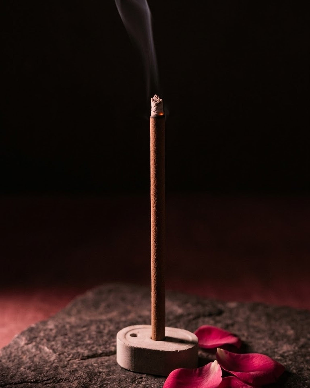 Glowing Gul Aangan dhoop stick in its holder with smoke rising, beside rose petals