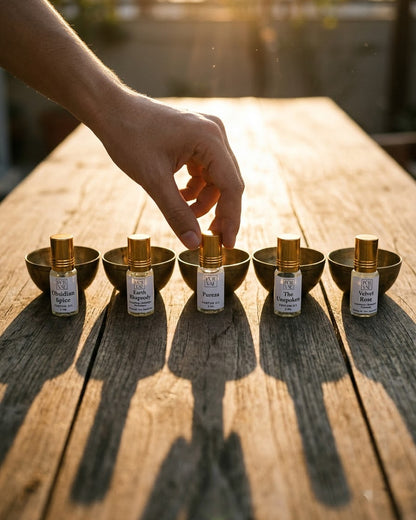 A hand reaching for one of five Purvae 3 ml parfum oil roll-ons standing in brass bowls on a wooden table at sunset