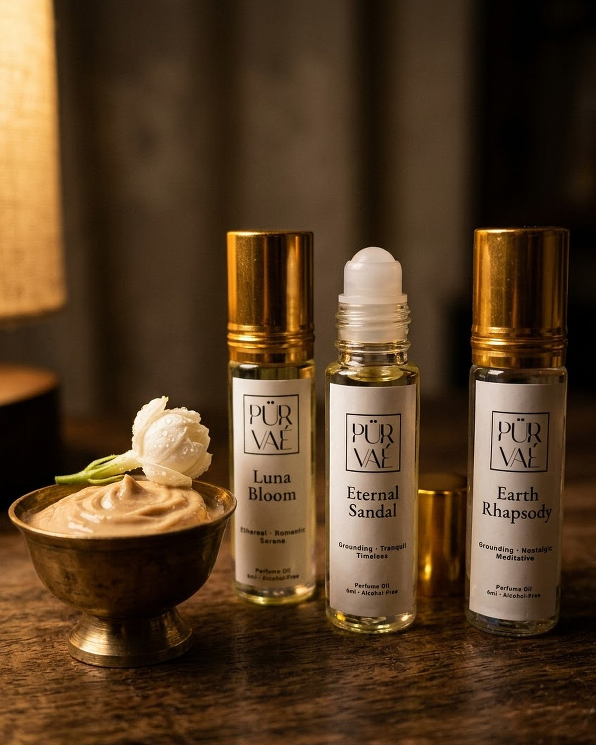 Earth Trio parfum oils beside a brass bowl of sandalwood paste topped with a jasmine bud, warm lamplight