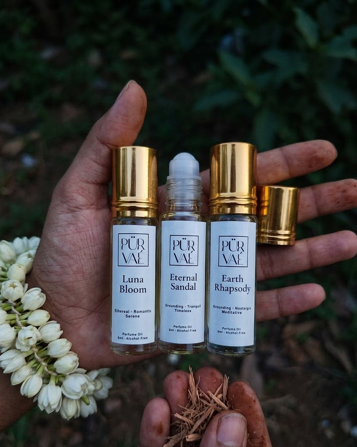 A hand holding the three 6 ml Earth Trio roll-ons with a jasmine garland and sandalwood shavings