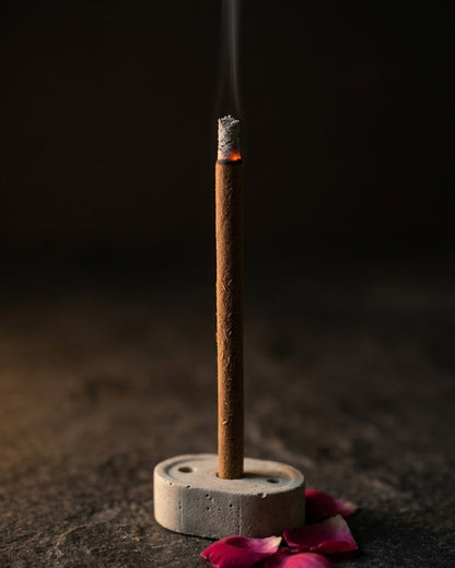 Close-up of a glowing Gul Aangan dhoop stick tip with a thread of smoke, beside rose petals