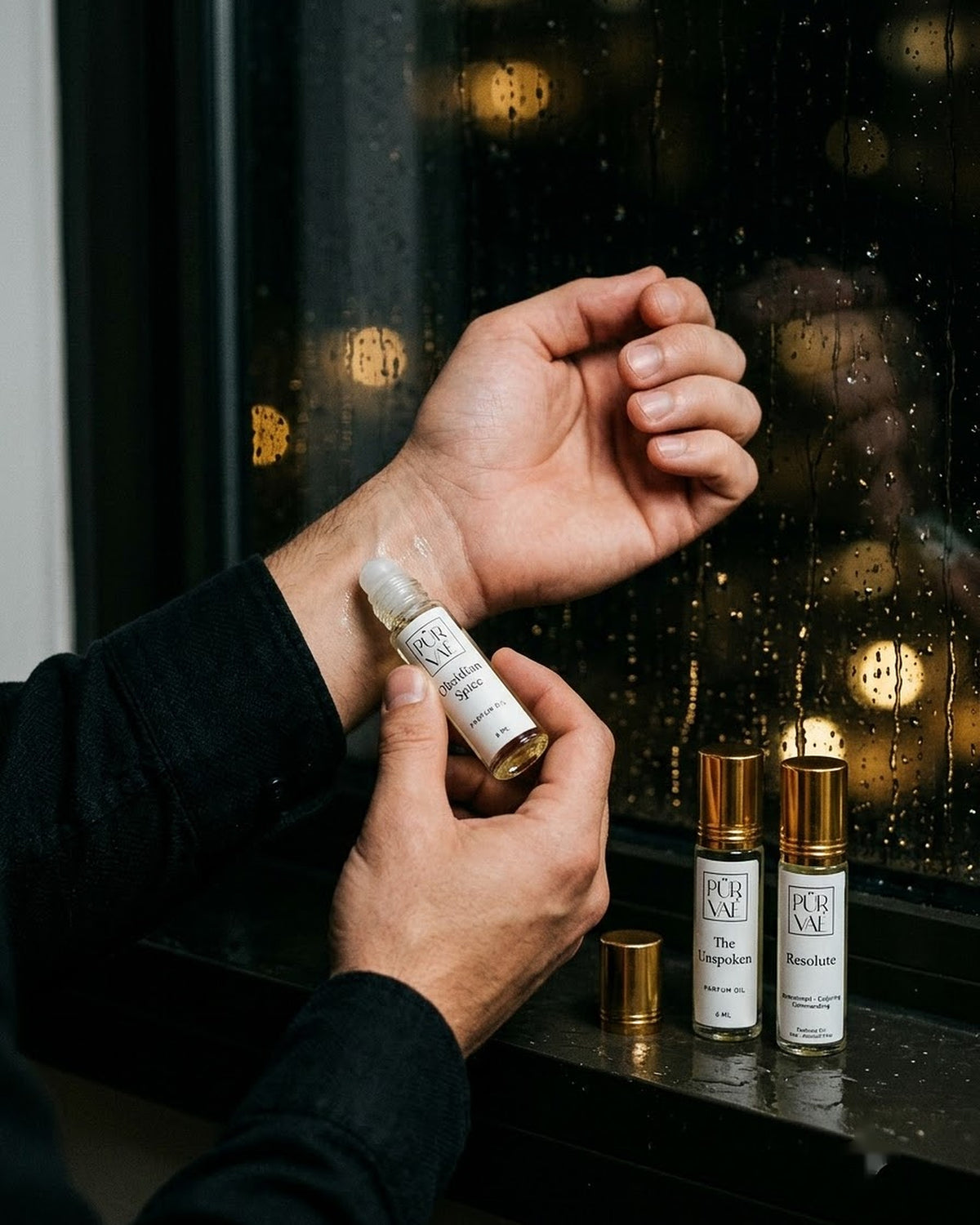 A man applying Obsidian Spice parfum oil to his wrist at a rainy window, the other two Night Trio roll-ons on the sill